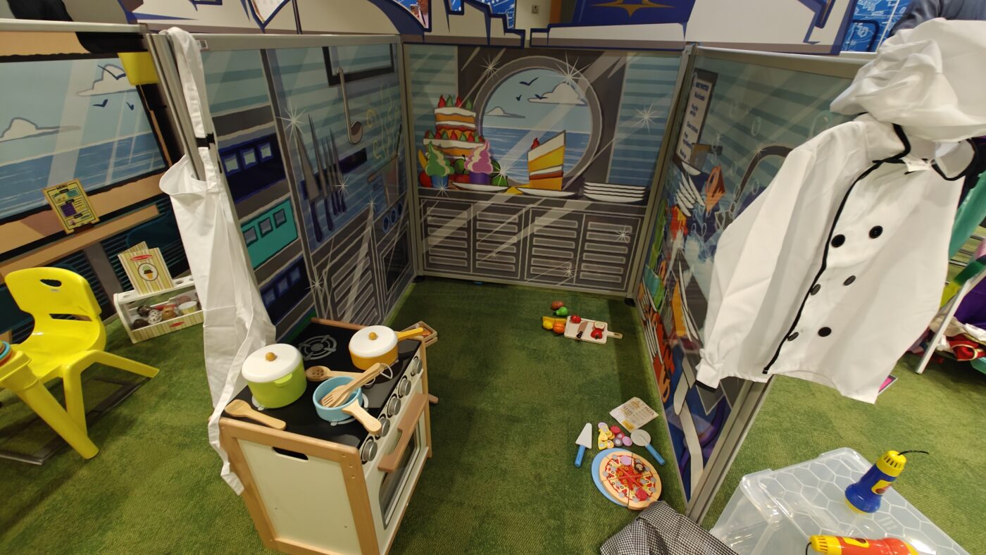 Modular galley play zone panel fully dressed with custom wooden food toys, cooker costume and props, and tactile toys.